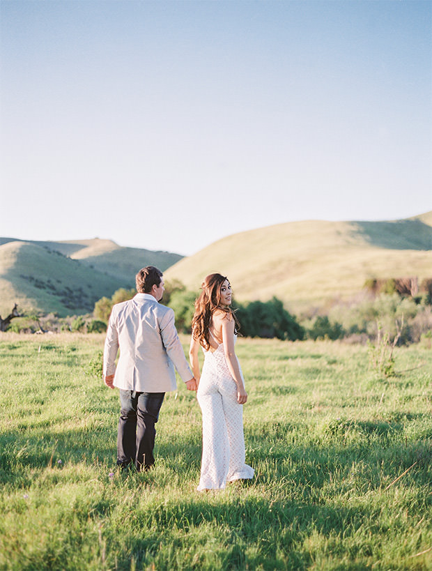 Woman in a white jump suit looks back while holding hands with her fiancé in rolling hills