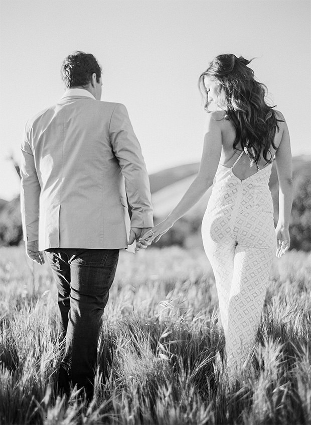 black and white image of a couple holding hands walking away in a grass field