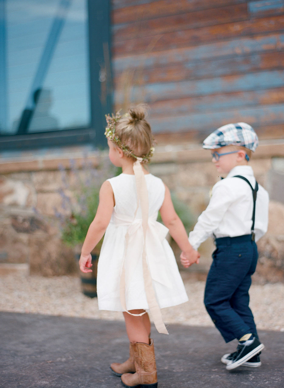 A flower girl with a flower crown and white dress holds the hand of a short ring bearer with blue glasses, suspenders and a checkered cap and walk away together. 