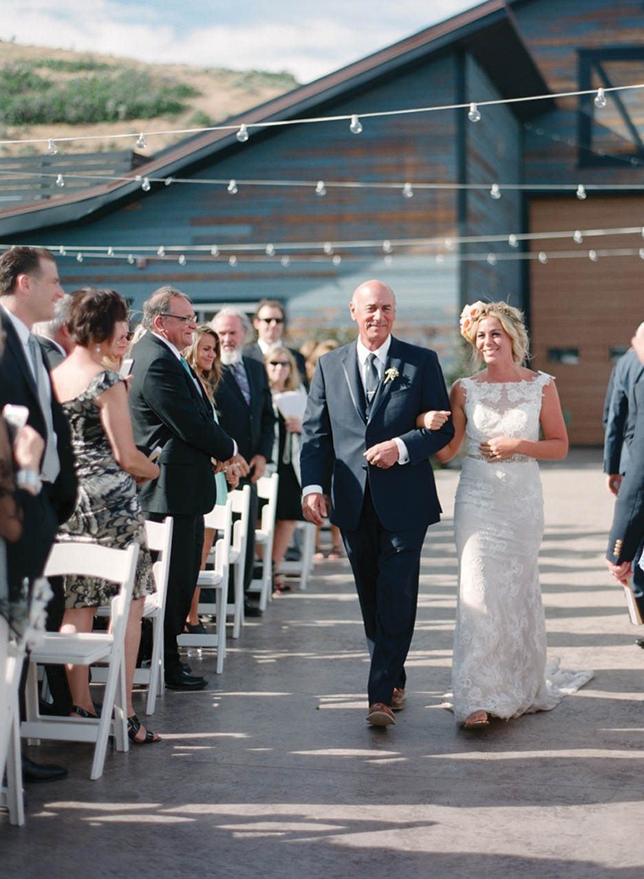 The bride walks down the aisle smiling with her father under string lights at High West Distillery.