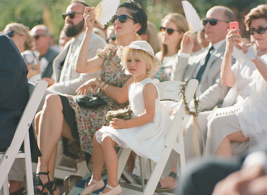 A view of weddings guests fanning themselves during a ceremony with the focal point on a flower girl with a hat in all white.