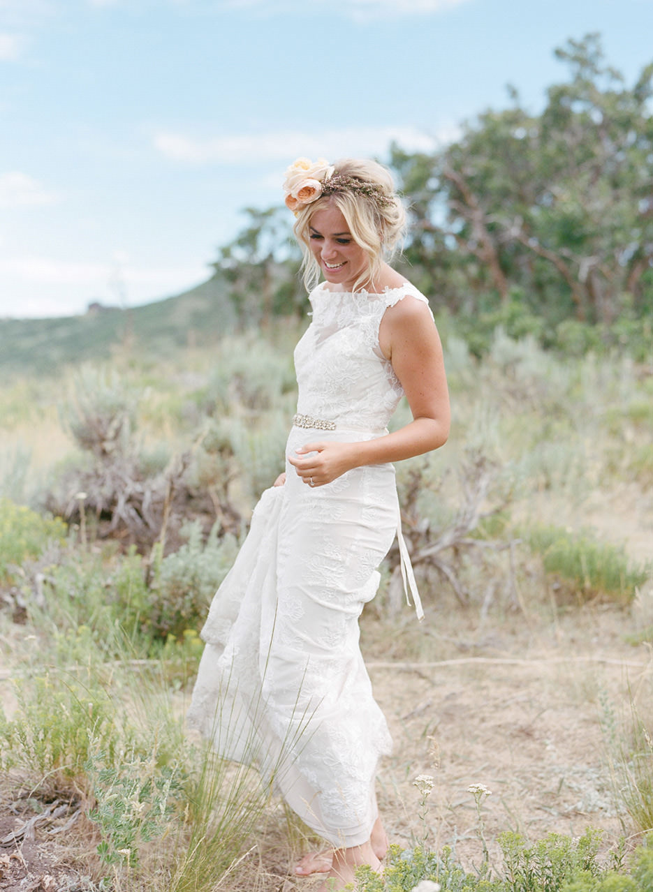 A barefoot bride with a flower crown smiles as she holds her dress with her right hand, stands in the blue sky ranch brush.