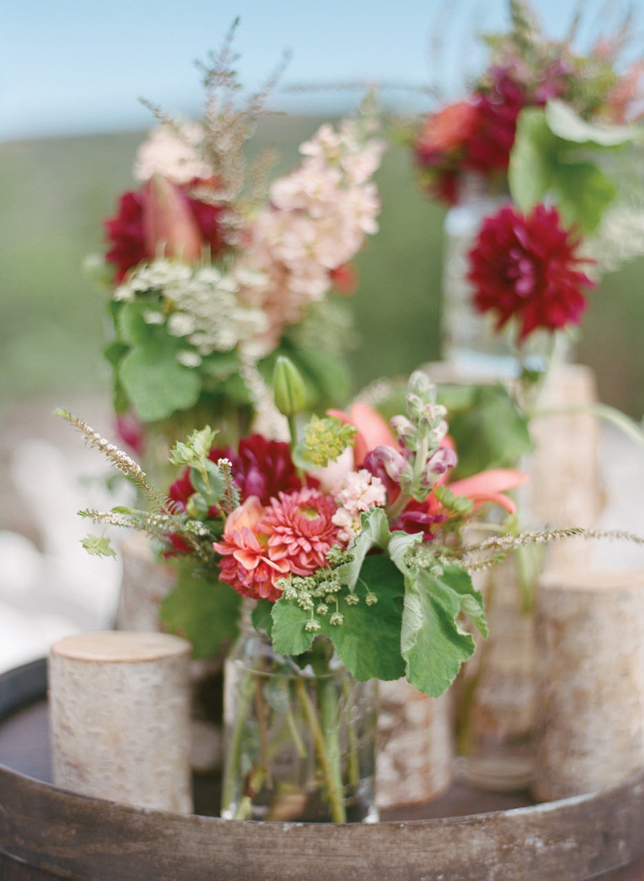 Floral arrangements in peach, dark magenta, and pink are held in mason jars with logs surrounding it. 