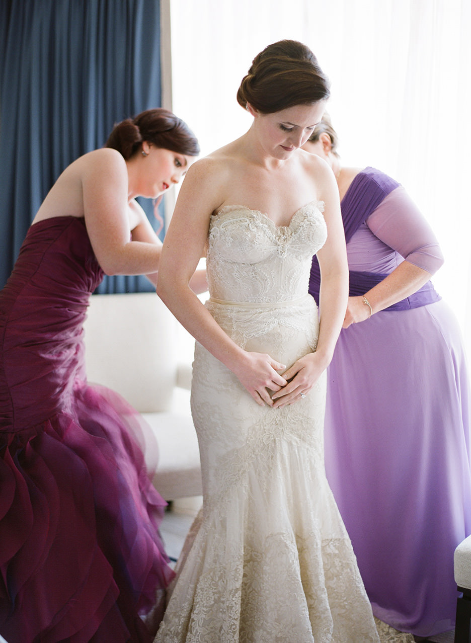 A bride in a cream strapless dress is being button up a bridesmaid in burgundy and mother in purple at the Peninsula Hotel Chicago for Wedding Day Tips and Tricks