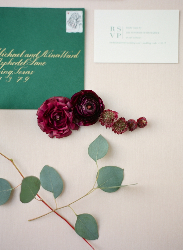 An invitation suite with berry colored flowers and eucalyptus leaves adorn a hunter green and gold invitation suite. 