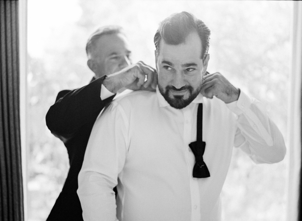 A black and white of a father helping his son (the groom) tie his bowtie. 