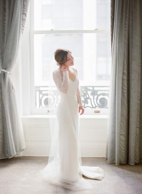 A bride in a long sleeve lace gown adjusts her earrings in front of the window of a room in the Palace Hotel in San Francisco. 