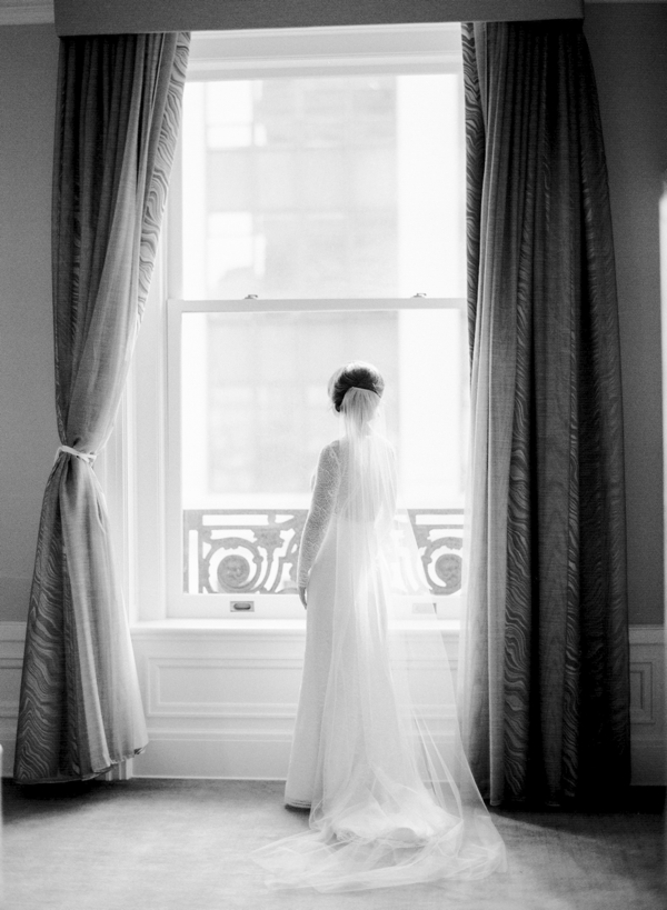 A bride in a long sleeve lace gown stands in front of the window of a room in the Palace Hotel in San Francisco. 