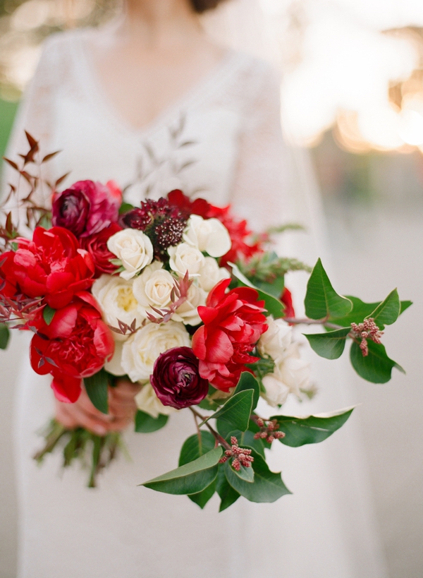A bouquet of deep red and cream ranunculus is held by a bride in a long sleeve lace gown. 