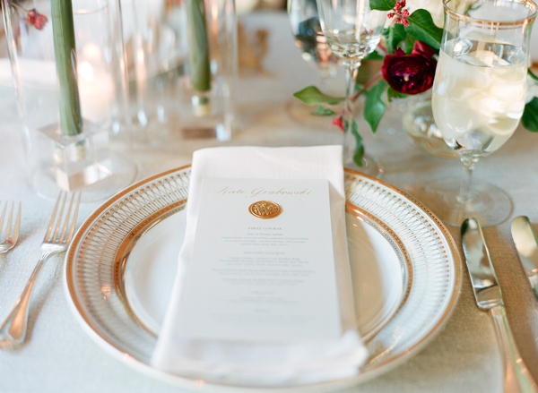 Cream roses and cranberries in small glass vases surrounded with candles adorn gold and white place setting complete with wax stamp on the menu. 