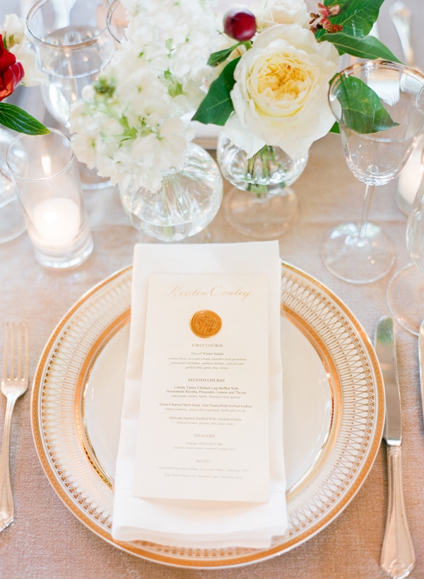Cream roses and cranberries in small glass vases surrounded with candles adorn gold and white place setting complete with wax stamp on the menu. 