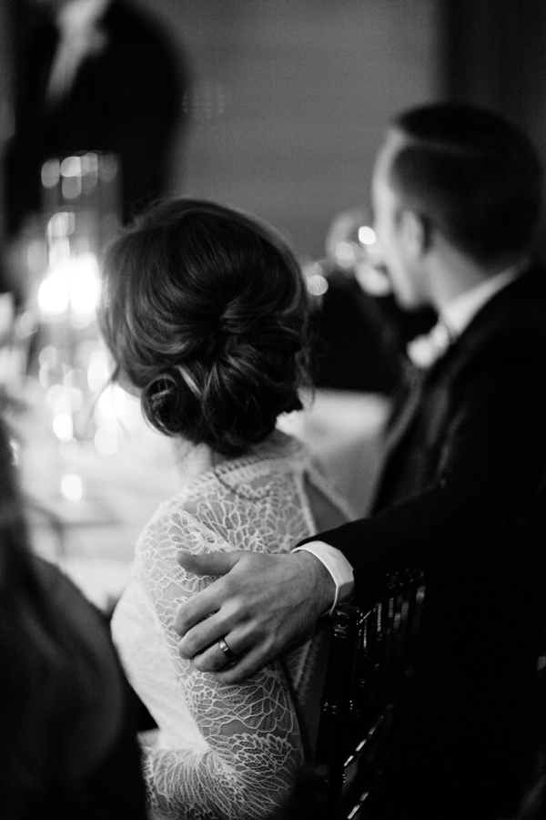 A groom puts his arm around his bride as they sit and listen to a speech by the best man. 