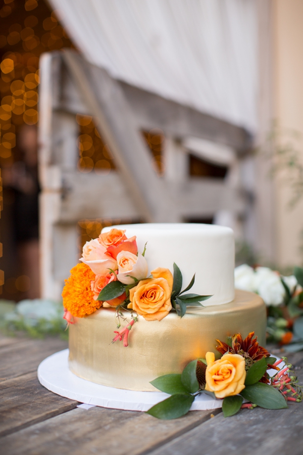 Small gold and white cake adorned with peach and orange roses in front of the barn at Holman Ranch. 