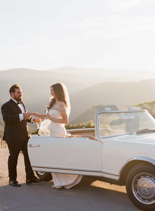 A bride and groom share a glass of champagne as she steps out of the getaway Mercedes convertible car. 