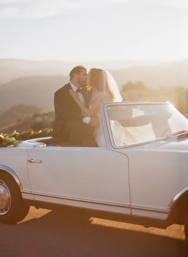 A bride and groom share a kiss as she steps out of the getaway Mercedes convertible car. 