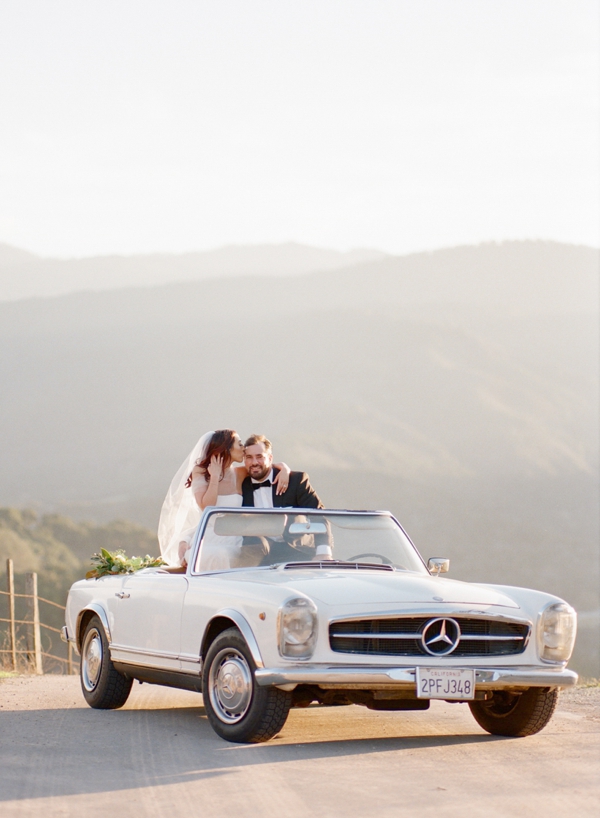 A bride and groom sit on top of their convertible getaway car and share a moment.