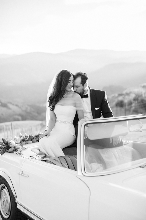 A bride and groom sit on top of their convertible getaway car and share a moment.