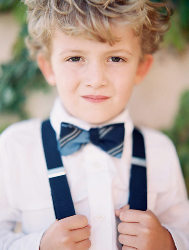 A blonde ring bearer holds his blue suspenders for a portrait.