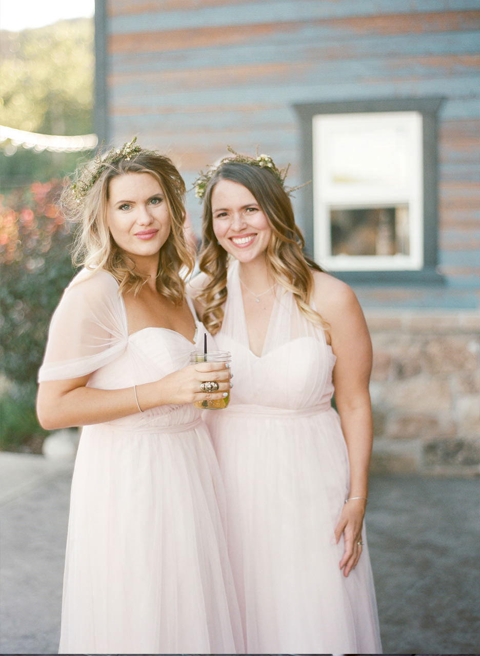 Two bridesmaids wearing flower crowns and light pink dress smile together.