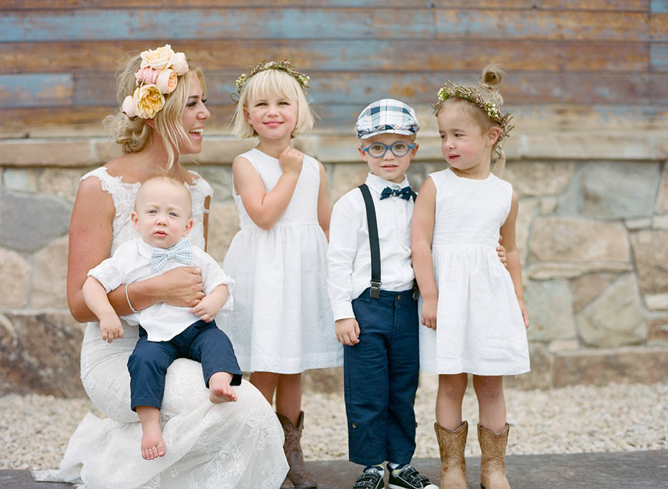 A bride with a flower crown share laughter with 2 flower girls wearing white dresses and two ring bearers wearing blue suspenders.