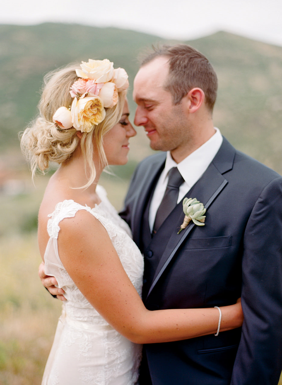 A bride with a yellow and pink flower crown cuddles with her arms around the groom.