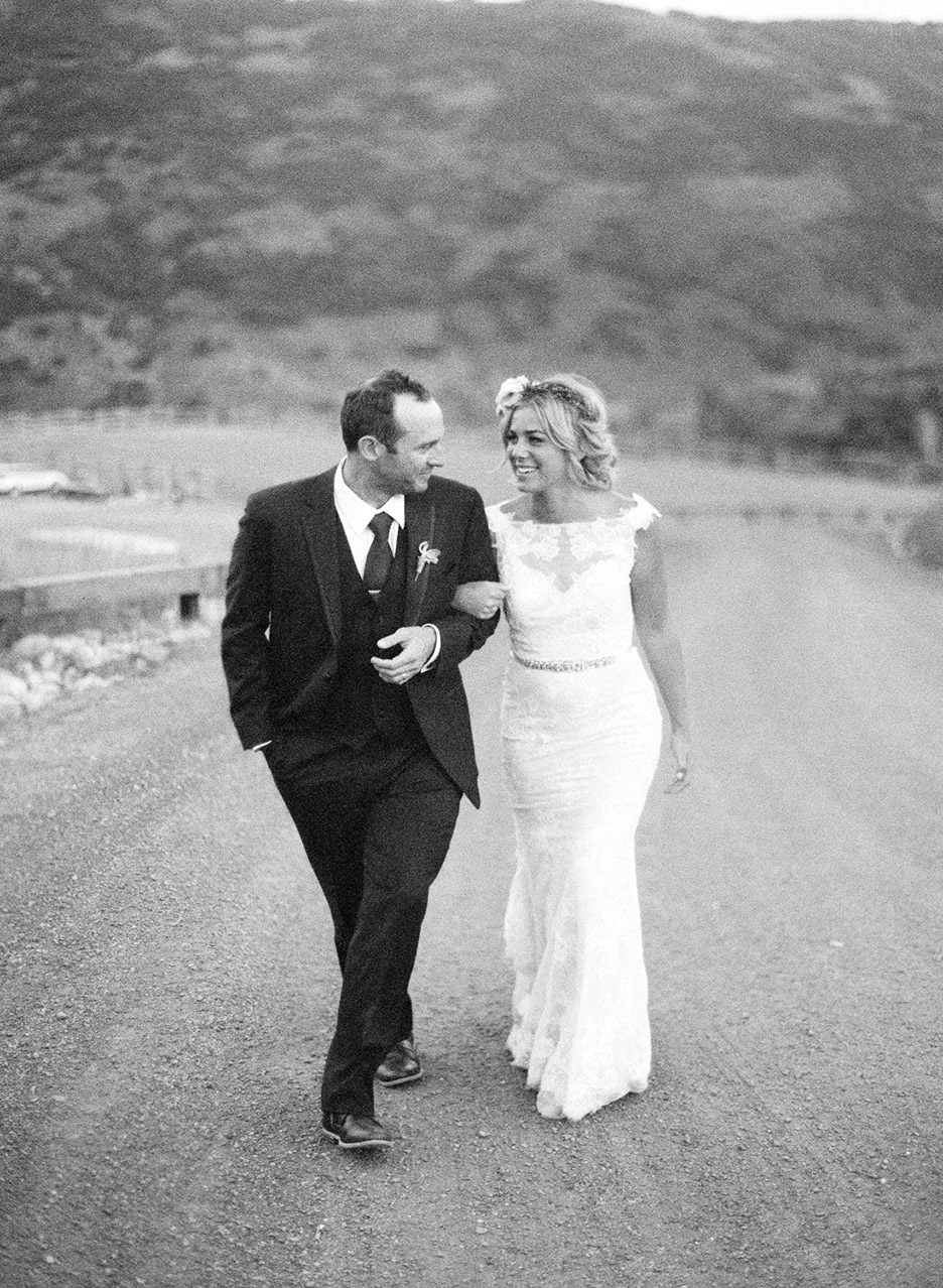 A bride and groom walk with linked arms up a gravel road in the mountains.