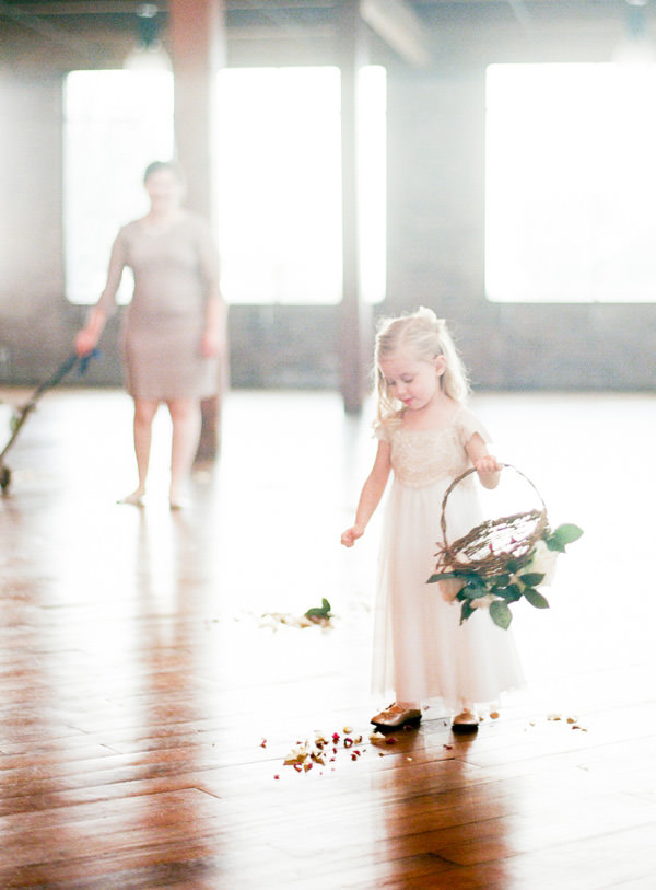 A blonde flower girl throws petals on to the wood floor from her basket as she walks down the aisle with her mother in the background.