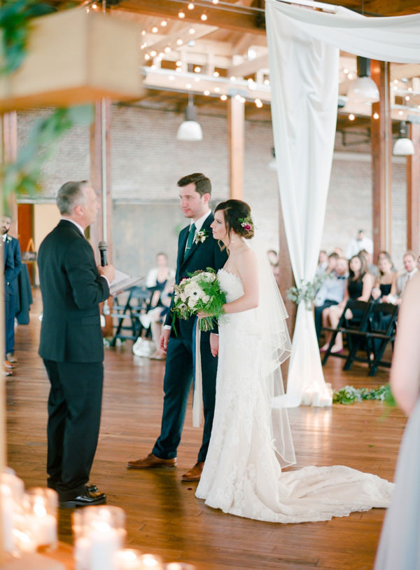 Bride and groom in an industrial venue with candles and drapes, stand beside each other and listen to the pastor speak to them.