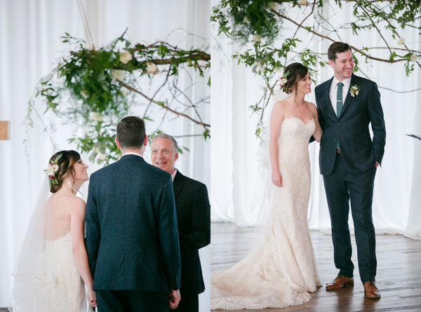 Bride and groom smile together during their ceremony in front of white drapes and a rose and green branch backdrop