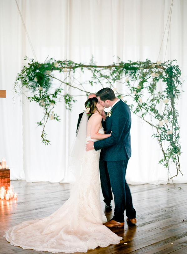Bride and groom exchange first kiss as husband and wife in front of an asymmetrical rose and branch backdrop