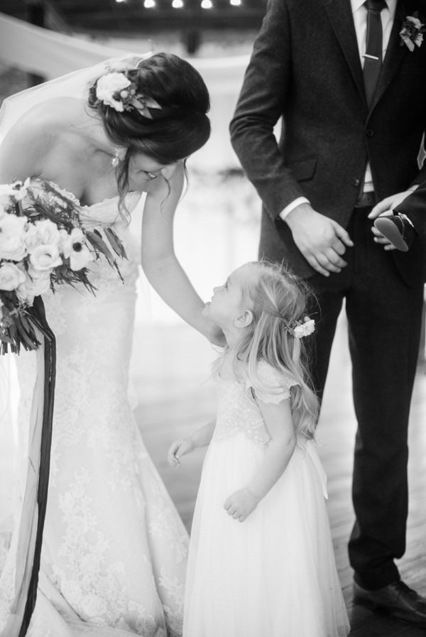 Bride holding flowers leans down and smiles at the flower girl who looks up happily at her (black and white)
