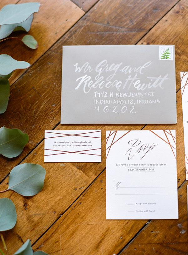 A grey and white hand calligraphy invitation suite address to Indianapolis, Indiana arranged on a wood floor.