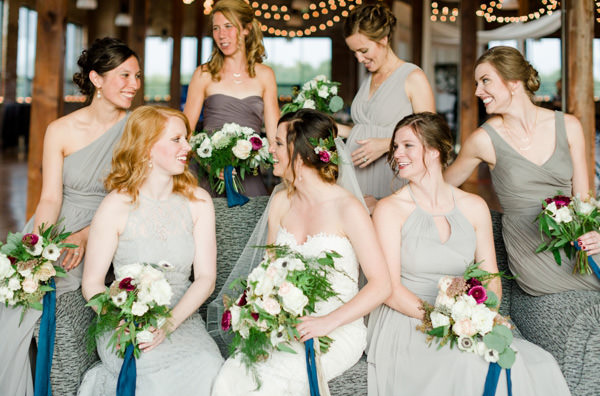 Bride and bridesmaids in grey dresses sit on the sofa with their bouquets laughing and smiling as they talk.