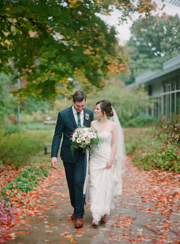 A bride holds her dress from the wet sidewalk strewn with autumn colored wet foliage walking besides her husband.