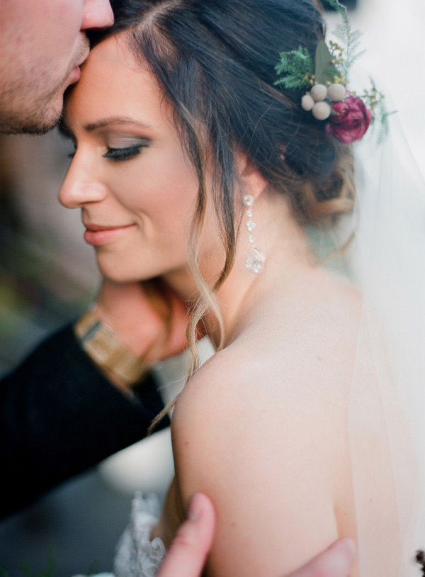 A bride with a small flower and berries in the side of her pinned up hair receives a sweet kiss from her husband on the forehead.