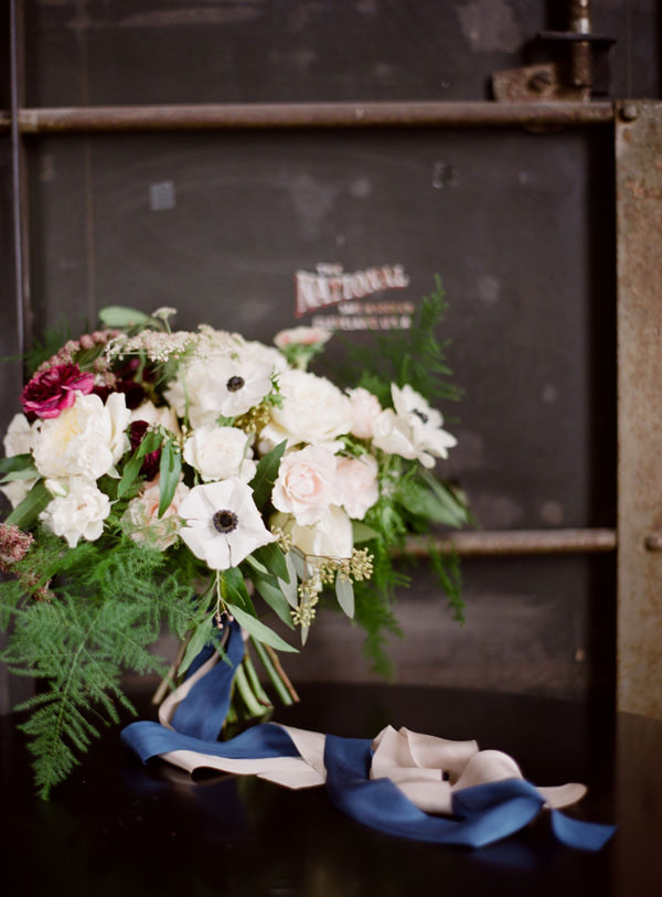 A white anemone and pink ranunculus bouquet with blue and blush silk ribbon sitting on a table in front of an industrial door.