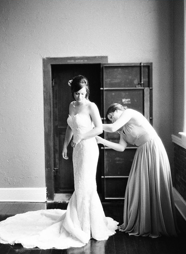Black and white Film image of a Maid of Honor zipping up her sister (the bride) in front of an industrial doorway. 