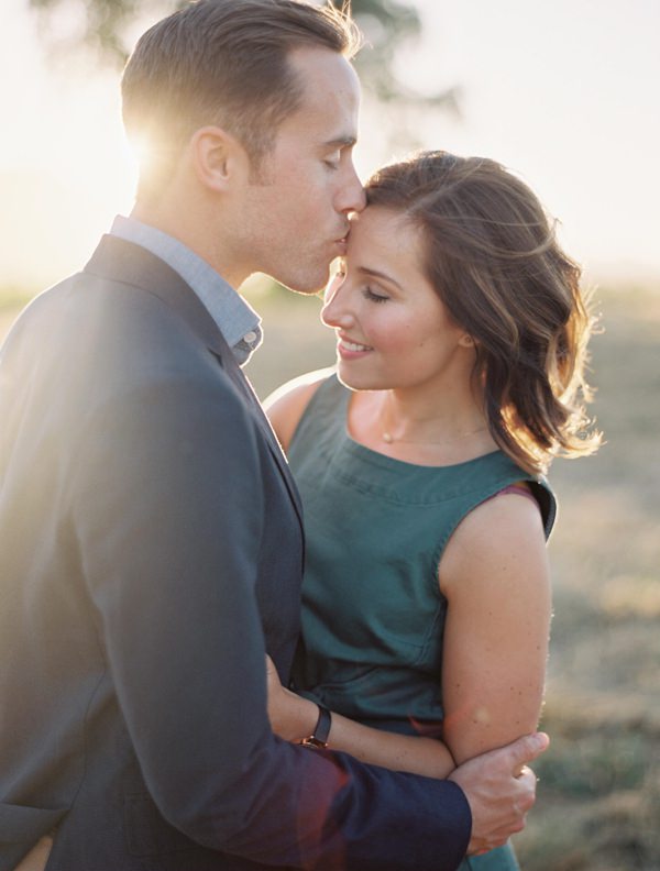 Smiling girl gets a kiss of the forehead from her fiancé while the sun sets