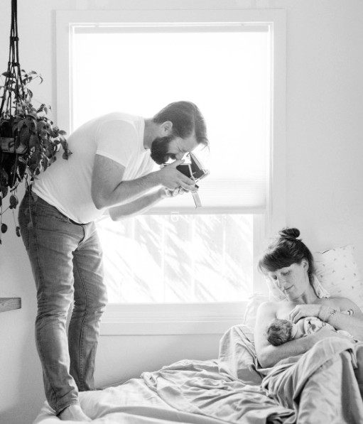 Father standing on the bed barefoot taking a Polaroid of his newborn son breastfeeding.